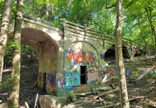 Abandoned concrete bridge covered in colorful graffiti, surrounded by lush green trees and underbrush.