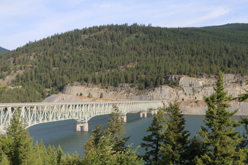 A steel bridge spans a river, surrounded by lush green hills and rocky cliffs under a clear blue sky.