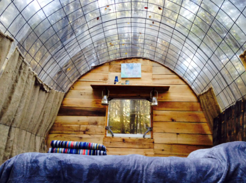 Interior of a cozy, rustic cabin with wooden walls, a large window, and a bed covered in blankets. Natural light filters in.