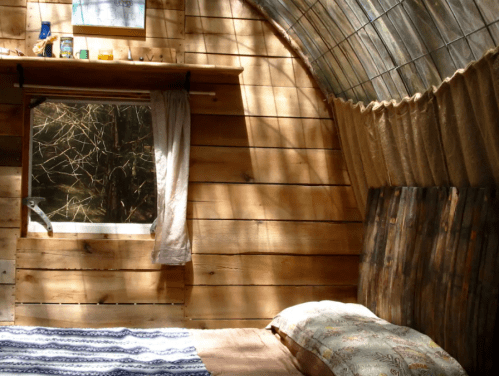 Cozy interior of a rustic cabin with wooden walls, a bed, and a window letting in natural light.