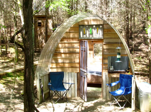 A cozy wooden cabin in a forest, featuring a curved roof, open door, and two blue chairs outside.