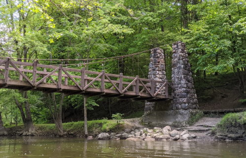 A wooden suspension bridge spans a river, flanked by stone pillars and surrounded by lush green trees.