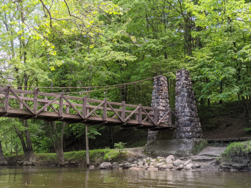 A wooden suspension bridge with stone supports over a calm river, surrounded by lush green trees.