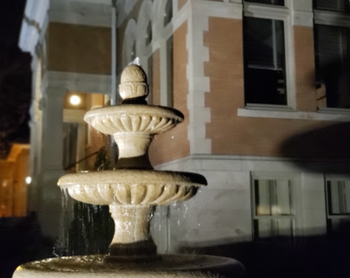 A stone fountain with three tiers, illuminated at night, in front of a historic building. Water gently flows from the top.