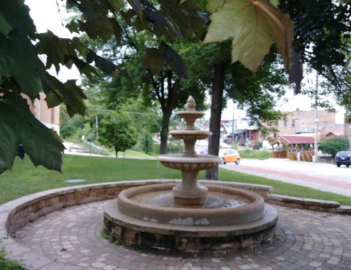 A stone fountain surrounded by greenery, with a view of a street and buildings in the background.
