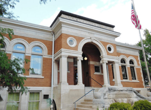 Historic brick building with arched entrance, steps, and an American flag in front. Green trees frame the scene.