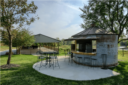 A rustic outdoor seating area with a round kiosk and a covered pavilion surrounded by greenery.