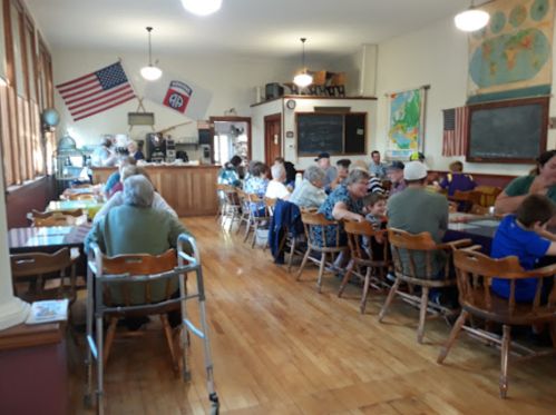 A busy dining hall with people seated at wooden tables, flags on the walls, and a counter in the background.