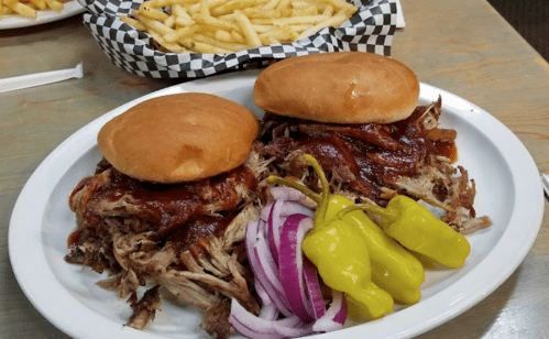 Two pulled pork sandwiches with barbecue sauce, served with sliced red onions and yellow peppers, alongside a basket of fries.