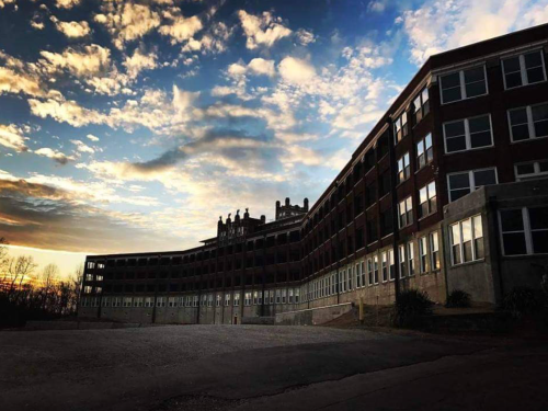 A historic building with a curved facade against a dramatic sunset sky, featuring clouds and a hint of trees in the background.