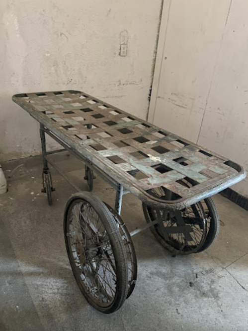 A vintage metal cart with a slatted top and two large wheels, positioned against a plain wall.