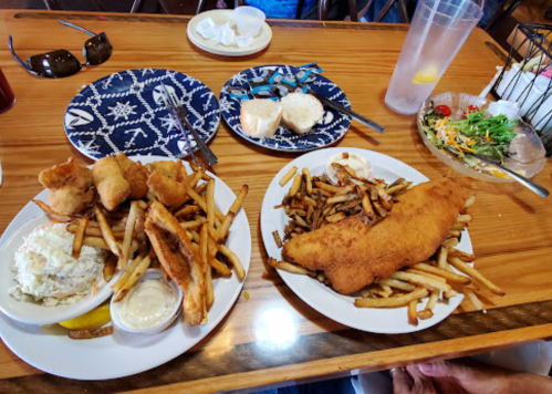Two plates of fried fish and fries, with coleslaw and dipping sauce, on a wooden table with drinks and bread.