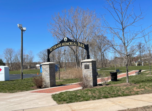 Entrance to Veterans Memorial Park, featuring a stone archway and clear blue sky. Green grass and trees in the background.