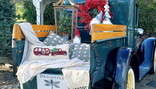 A vintage truck bed decorated with festive pillows, a lantern, and holiday accents, surrounded by greenery.