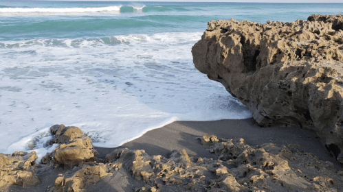 Waves gently lap against rocky shore, with clear blue water and sandy beach in the background.