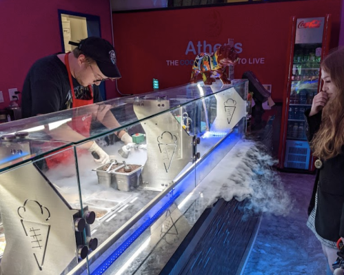 A person prepares ice cream at a counter, with smoke effects and a customer observing in a colorful shop.