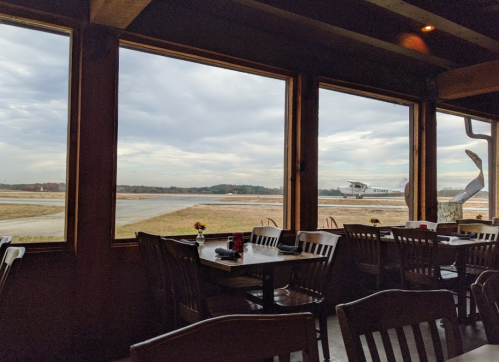 View from inside a restaurant with wooden tables, looking out at a small airplane on a runway under a cloudy sky.