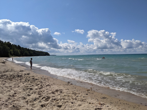 A serene beach scene with a person standing on the shore, gentle waves, and a boat in the distance under a cloudy sky.