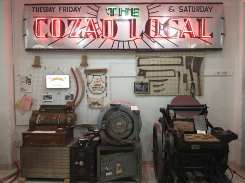 Neon sign reading "The Cozad Local" with vintage printing equipment and a cash register displayed on a wall.
