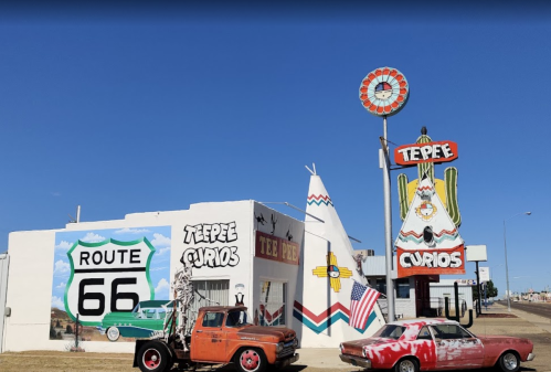 Colorful roadside attraction featuring a teepee, vintage cars, and a Route 66 sign against a clear blue sky.