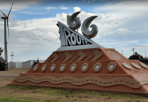 A colorful Route 66 sign with a wave design, set against a cloudy sky and wind turbines in the background.