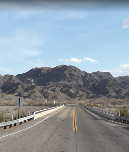 A deserted road stretches towards mountains under a clear blue sky, with sparse vegetation on either side.