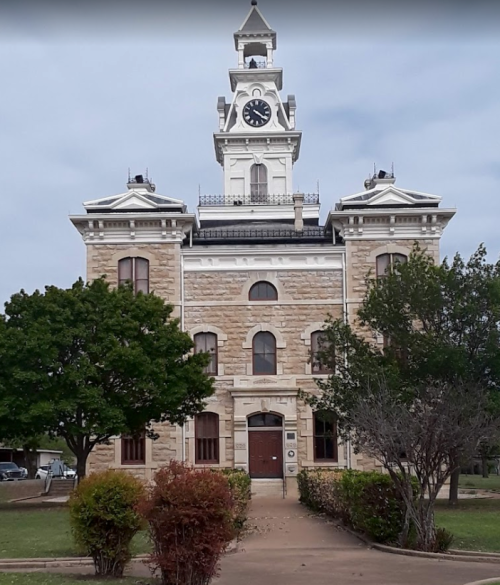 Historic stone building with a clock tower, surrounded by trees and a pathway, under a cloudy sky.