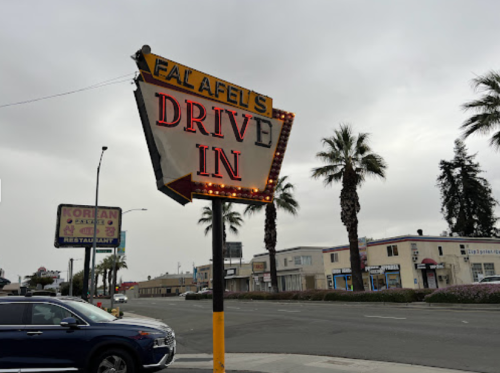 A retro sign for "Falafel's Drive In" with palm trees and a cloudy sky in the background.