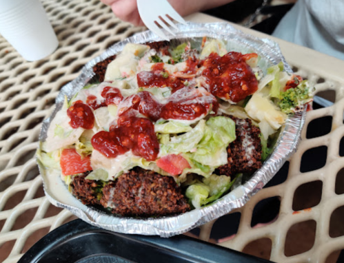 A foil bowl filled with salad, crispy falafel, and drizzled with a red sauce, placed on a textured surface.