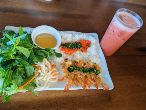 A plate of shrimp with rice noodles, fresh herbs, pickled vegetables, and a dipping sauce, alongside a pink beverage.