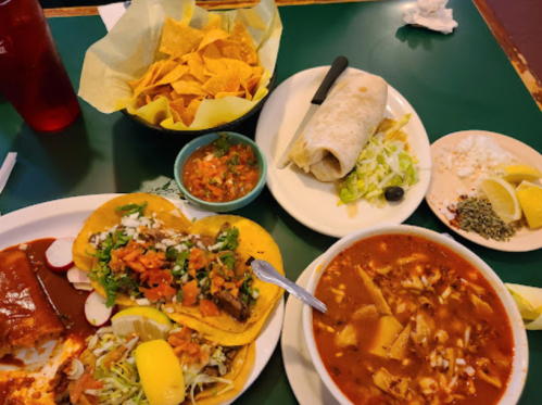 A table spread with tacos, a burrito, tortilla chips, salsa, and a bowl of soup, garnished with lemon and herbs.