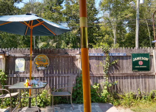 Outdoor dining area with a table, umbrella, and a wooden fence featuring various signs and greenery.