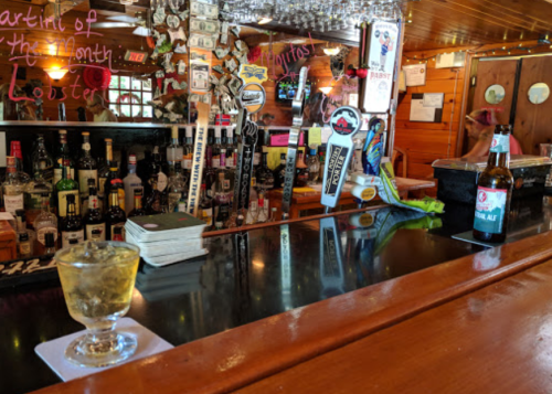 A cozy bar scene featuring a drink on the counter, a variety of bottles behind, and a warm wooden interior.