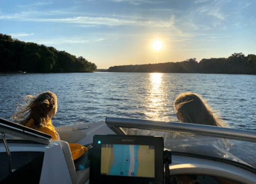 Two girls sit on a boat, watching the sunset over a calm river, with a navigation screen in the foreground.