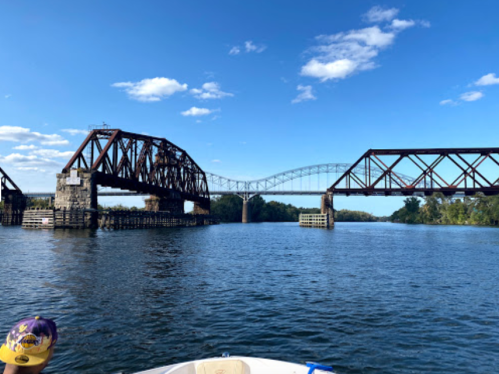 A view of two bridges over a river, with blue skies and trees in the background, seen from a boat.