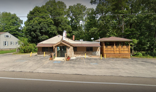 A single-story building with a stone facade, surrounded by trees, featuring a porch and parking area.