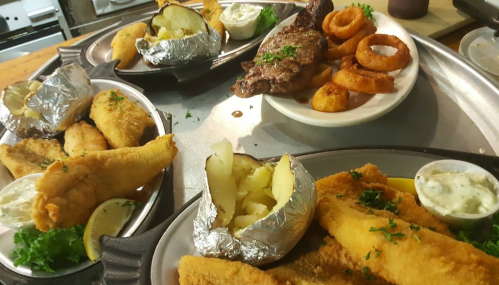 A tray of assorted dishes including fried fish, steak, baked potatoes, onion rings, and dipping sauces.