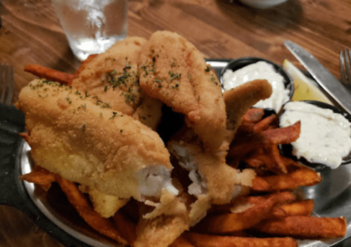 Fried fish fillets on a bed of sweet potato fries, served with dipping sauces and a lemon wedge.