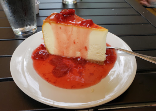 A slice of cheesecake topped with strawberry sauce on a white plate, with a glass of water in the background.