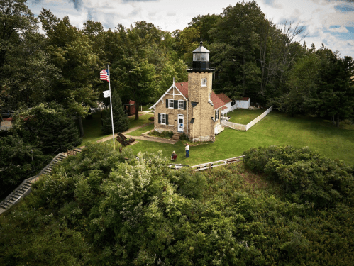Aerial view of a lighthouse surrounded by greenery, with a flag flying and two people near the building.