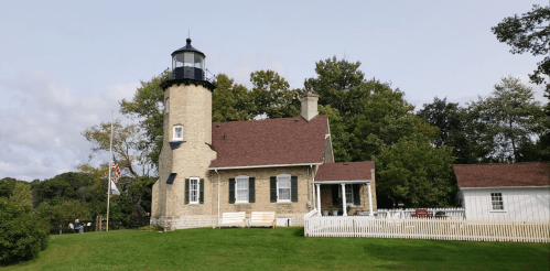 A historic lighthouse with a stone tower and a red-roofed house, surrounded by green grass and trees.