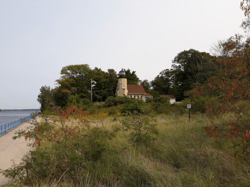 A coastal scene featuring a house surrounded by trees and tall grass, with a pathway and water in the background.