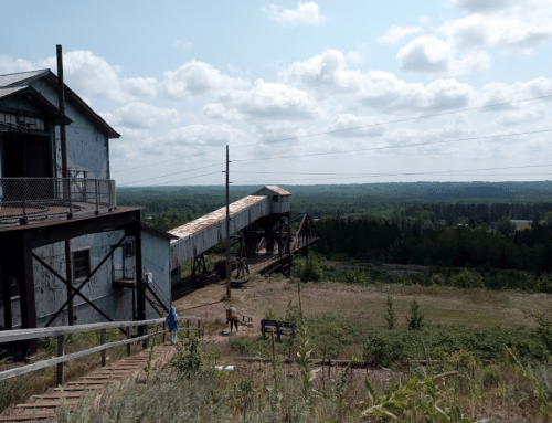 An old industrial building with a conveyor belt extending over a green landscape under a cloudy sky.