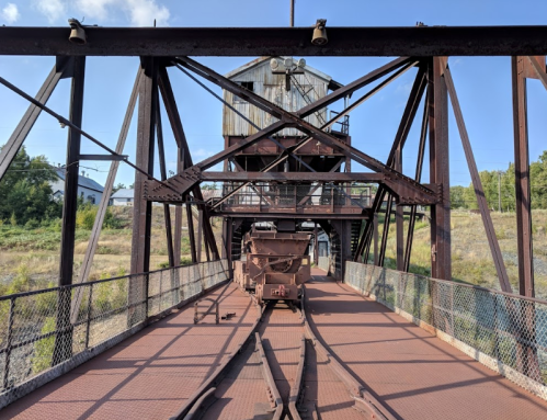 A rusted industrial structure with tracks leading to a loading area, surrounded by greenery and blue sky.
