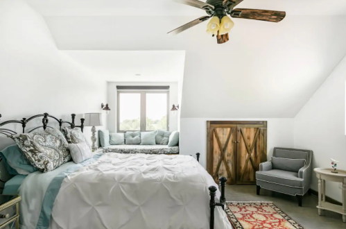 Cozy bedroom with a large bed, decorative pillows, a seating area, and rustic wooden doors. Bright and airy atmosphere.