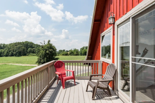 A red barn with a wooden deck featuring two chairs, overlooking a green field under a partly cloudy sky.