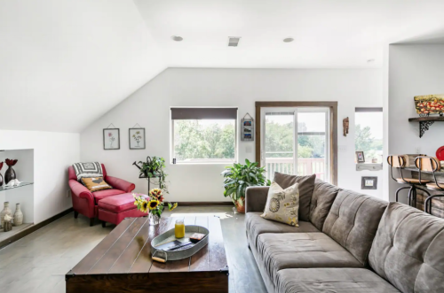 Cozy living room with a gray sofa, red armchair, plants, and a wooden coffee table, featuring large windows and natural light.