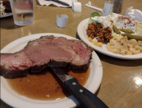 A plate with a large slice of roast beef in gravy, alongside a side of macaroni, pickles, and a meat dish.