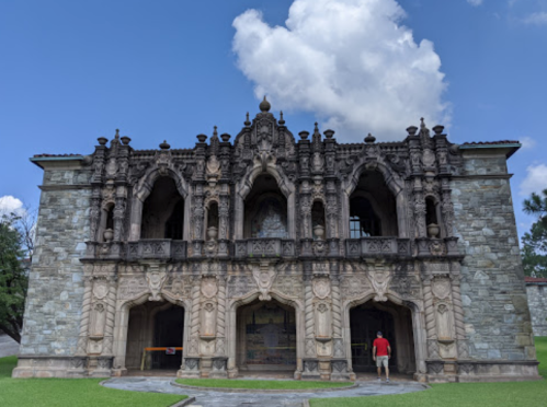 A stone building with intricate carvings and arches, set against a blue sky with fluffy clouds. A person stands nearby.