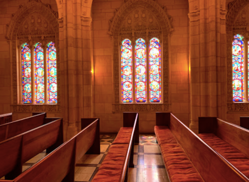 Interior of a church featuring wooden pews and colorful stained glass windows. Soft light illuminates the space.